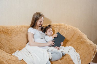 Hamile bir anne, küçük kızıyla kanepede oturuyor ve bir aletle oynuyor.