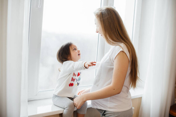 A pregnant mother is standing near the window with her little daughter