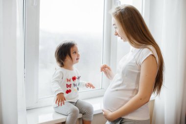 Hamile bir anne, küçük kızıyla pencerenin yanında duruyor.