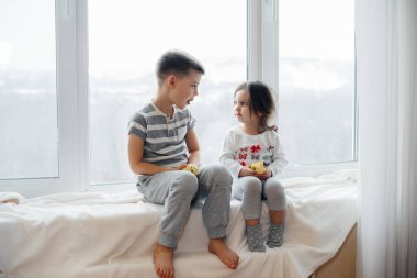 Erkek ve kız kardeş pencere kenarında oturuyor ve elma yiyorlar. Mutluluk