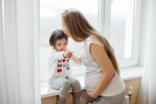 A pregnant mother is standing near the window with her little daughter