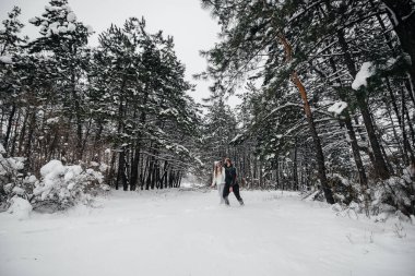 Bir çift, karlı bir ormanda, arka plandaki ağaçlara sarılıyor.