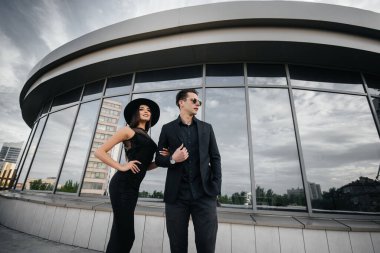 Siyah elbiseli ve gözlüklü güzel, şık bir çift genç gün batımında bir ofis binasının arka planında duruyor. Moda ve tarz