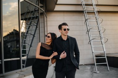 Siyah elbiseli ve gözlüklü güzel, şık bir çift genç gün batımında bir ofis binasının arka planında duruyor. Moda ve tarz