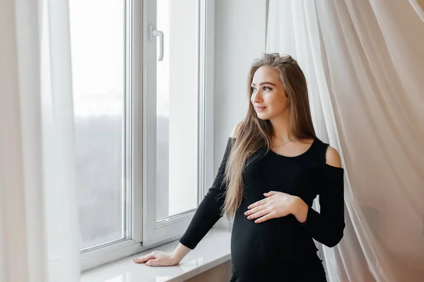 一个漂亮的怀孕女孩站在窗边的一个房间里 — 图库照片