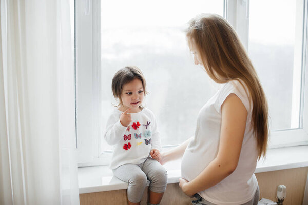 A pregnant mother is standing near the window with her little daughter