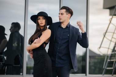 Siyah elbiseli ve gözlüklü güzel, şık bir çift genç gün batımında bir ofis binasının arka planında duruyor. Moda ve tarz