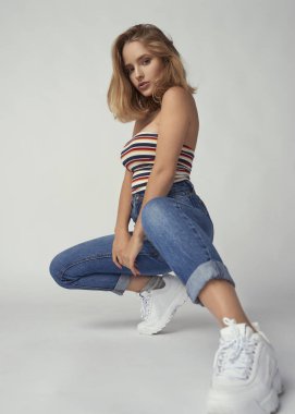 Çömelmiş genç ve atletik bir kız kot pantolon ve kısa tişört, güzel ve sağlıklı ten, doğal sarı saç, boş arka plan, hassas makyaj