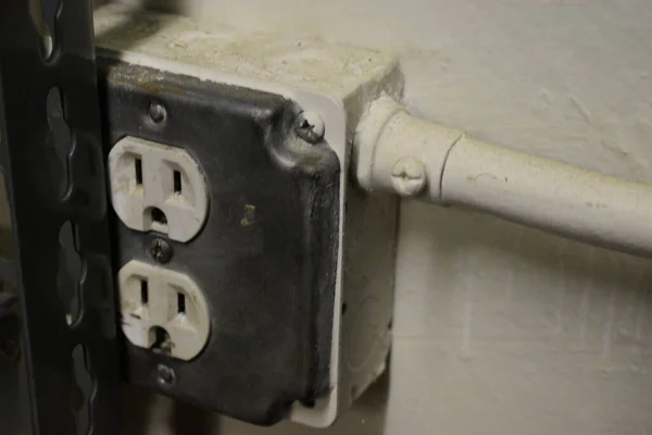 Gri metal, beyaz plastik, beyaz boru, beyaz duvar ve yakındaki metal raf bacağıyla kaplanmış elektrik prizi.. 