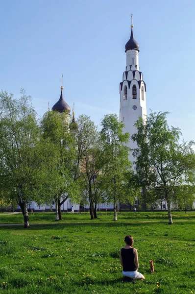 Молодая женщина с маленькой собачкой сидит на траве в парке и церковь на заднем плане — стоковое фото