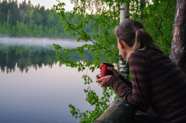 Isınmak için kahve ya da çay içme güzel genç kız. Portre çekici kadın düşünceli Gölü su yansıma ve holding üzerinden bakıyor.