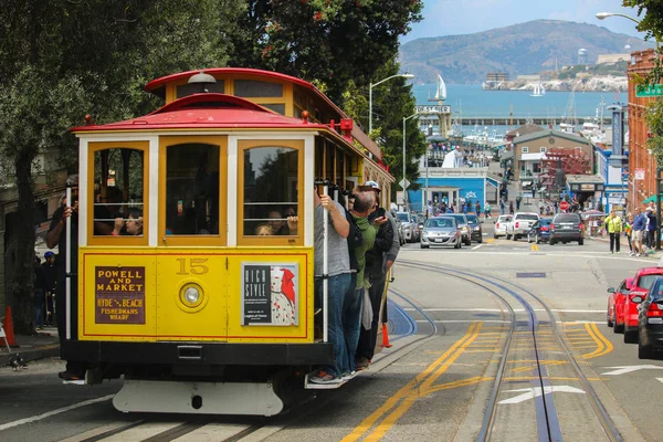 San Francisco, California - 23 de mayo de 2015: Turistas montados en el icónico teleférico, día de cielo azul en la cima de Hyde Street con vistas al agua de la bahía y la prisión de Alcatraz — Foto de Stock