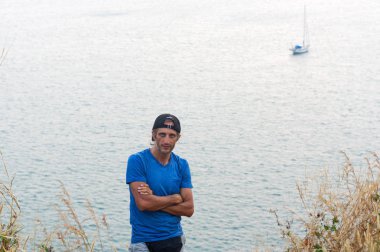 Mutlu genç sakallı adam ayakta bakış boş mavi tişört ve siyah kap giyen bir deniz manzarası ile