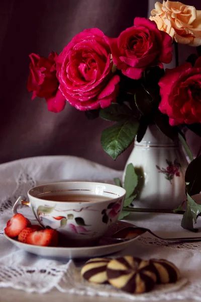 Una taza de té y galletas contra un jarrón con un ramo de rosas . — Foto de Stock