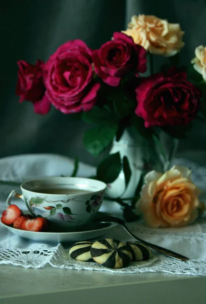A cup of tea and biscuits against a vase with a bouquet of roses. — Stock Photo, Image