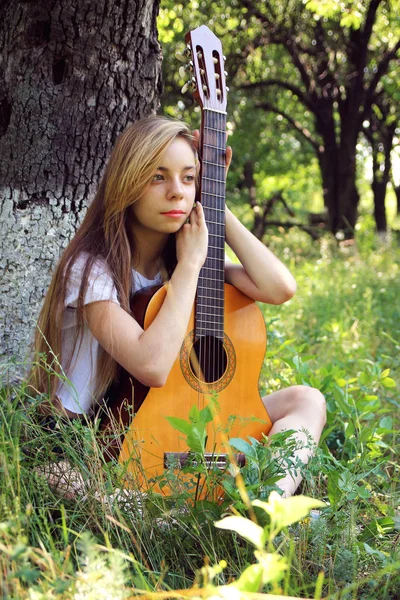 Den vackra flickan sitter eftertänksamt, lutande på en gitarr mot bakgrund av naturen. Stockbild