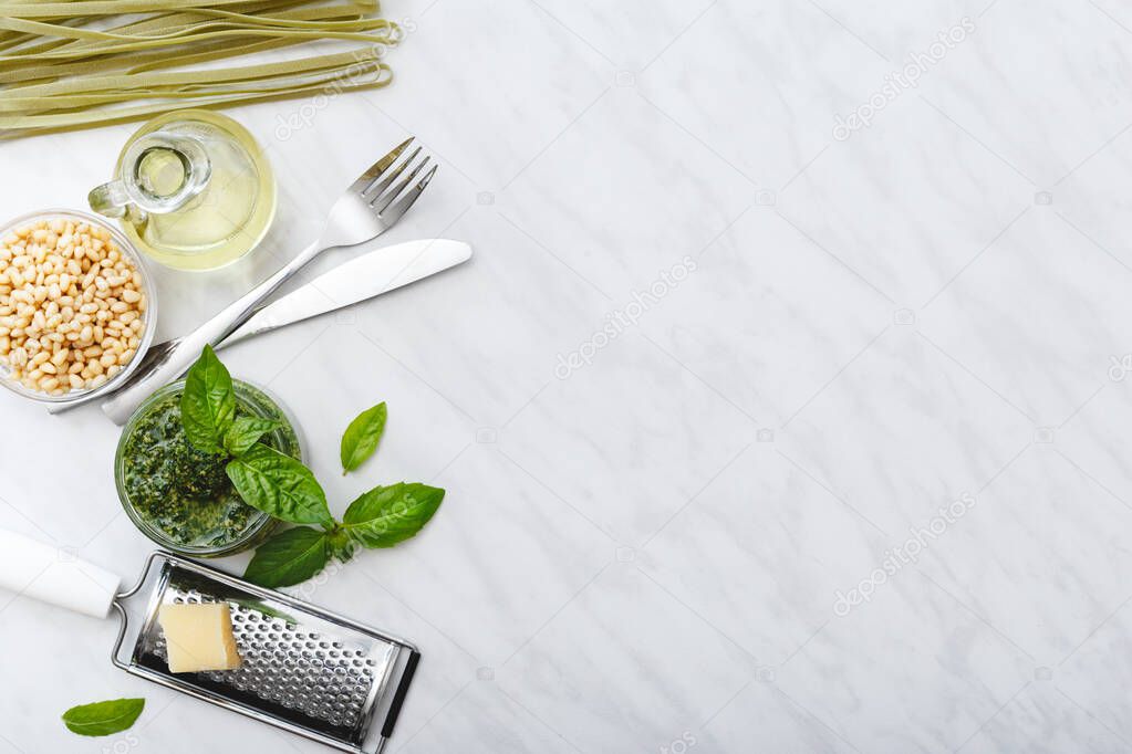 Raw green fettuccine and Pesto sauce with ingredients: pine nuts, basil, olive oil, parmesan and cutlery on white marble table. Making Pesto fettuccine. Top view, flat lay with copy space for text