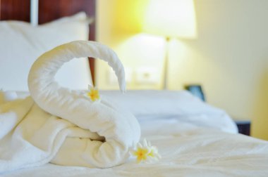 Beyaz banyo havlusunun güzel kuğusu beyaz yatakta süslenmiş. Havlu kuğusu ve topikal çiçekler - frangipani. Otelden güzel bir karşılama..
