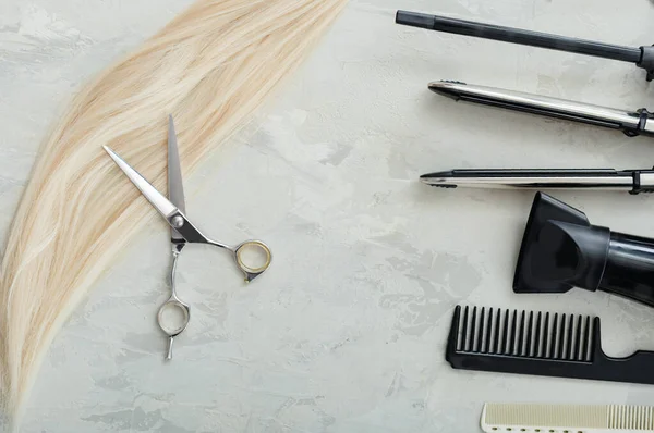 Platte lay compositie met kappers, schaar en draad van blond haar: schaar, kammen, strijkijzer op grijze achtergrond. Kapsalon service. Schoonheidssalon service. Kappersset. — Stockfoto