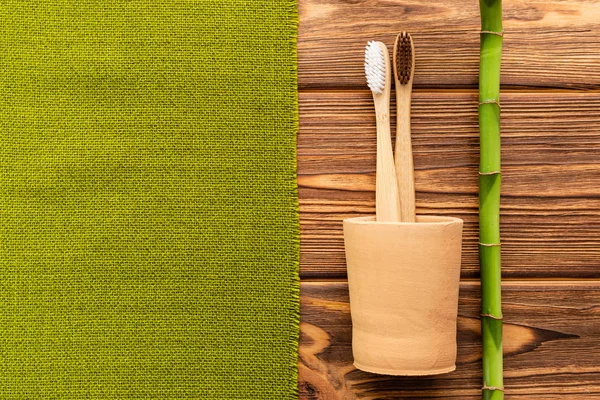 Bamboo toothbrushes in hand made clay glass bamboo plant with green towel on wooden background.Flat lay copy space. Biodegradable natural bamboo toothbrush. Eco friendly, Zero waste, Dental care — Stock Photo, Image