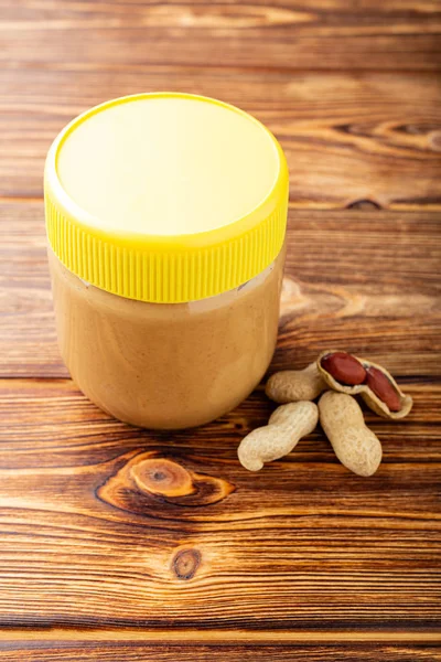 Creamy peanut paste in glass jar with yellow cap and peanuts in the peel scattered on the brown wooden table for cooking breakfast. Vegan food concept.