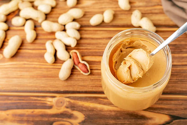 Cremige Erdnusspaste im offenen Glas, Erdnussbutter im Löffel. Erdnüsse in der Schale verstreut auf dem braunen Holztisch mit Kopierraum zum Kochen des Frühstücks. Veganes Ernährungskonzept. — Stockfoto