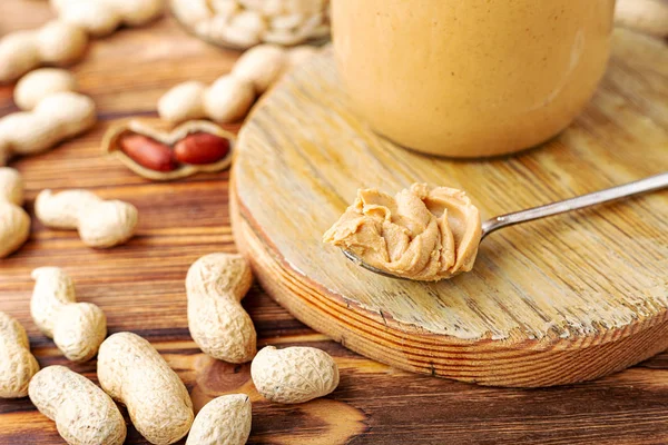 Mantequilla de maní en cuchara cerca de pasta de maní cremosa en frasco de vidrio abierto. Cacahuetes en la cáscara dispersos en la mesa de madera marrón con espacio de copia para cocinar el desayuno. Concepto de comida vegana . — Foto de Stock