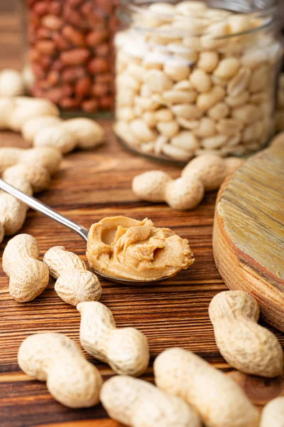 Mantequilla de maní en cuchara cerca pelada y cacahuetes en cáscara en frascos. Cacahuetes en la cáscara dispersos en la mesa de madera marrón con espacio de copia para cocinar el desayuno. Concepto de comida vegana . — Foto de Stock