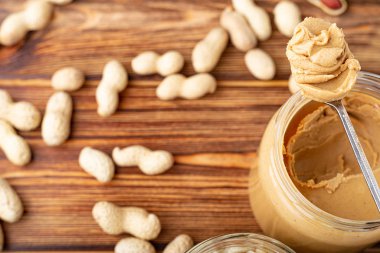 Açık cam kavanozda kremalı fıstık ezmesi, kaşıkta fıstık ezmesi. Kabuktaki fıstıklar kahverengi ahşap masaya saçılmış. Kahvaltı hazırlamak için de fotokopi alanı var. Vejetaryen gıda konsepti.