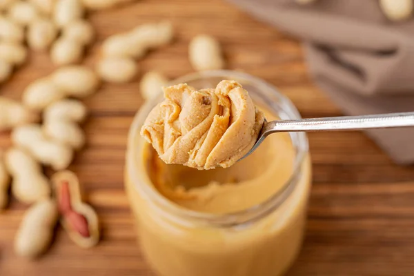 Erdnussbutter in einem Löffel in der Nähe von cremiger Erdnusspaste in einem offenen Glas. Erdnüsse in der Schale verstreut auf dem braunen Holztisch mit Kopierraum zum Kochen des Frühstücks. Veganes Ernährungskonzept. — Stockfoto