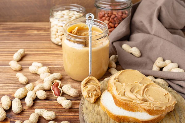 Erdnussbutter in einem Löffel neben cremiger Erdnusspaste im offenen Glas, Scheibe Erdnussbutter-Brot, Toast. Erdnüsse in der Schale verstreut auf dem braunen Holztisch mit Kopierraum zum Kochen des Frühstücks — Stockfoto