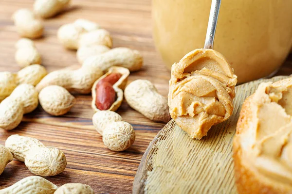 Peanut butter in spoon near creamy peanut paste in open glass jar, slice of peanut butter bread, toast. Peanuts in the peel scattered on the brown wooden table with copy space for cooking breakfast — 스톡 사진