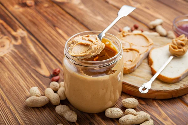 Erdnussbutter in einem Löffel neben cremiger Erdnusspaste im offenen Glas, Scheibe Erdnussbutter-Brot, Toast. Erdnüsse in der Schale verstreut auf dem braunen Holztisch mit Kopierraum zum Kochen des Frühstücks — Stockfoto