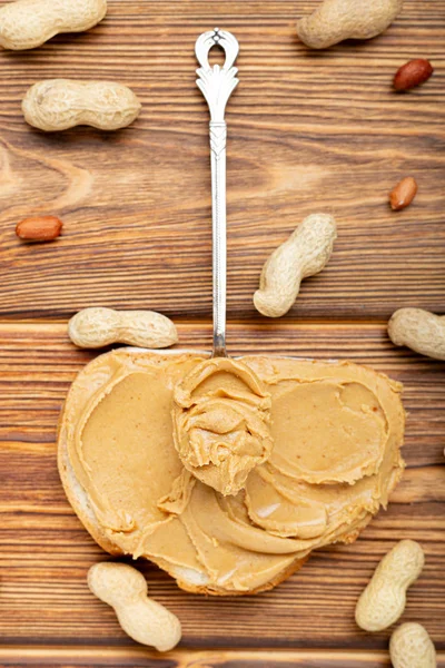 Vintage spoon with creamy peanut butter on a peanut butter bread. Peanuts in the shell and peeled peanuts on brown wooden table. Flat lay of peanut paste for cooking breakfast.Vegan food concept