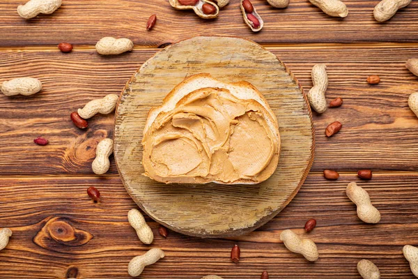 Sandwich de pan tostado con mantequilla de maní. Pasta de maní cremosa en un pedazo de pan. Cacahuetes en la cáscara dispersos en la mesa de madera marrón con espacio de copia para cocinar el desayuno. Concepto de comida vegana — Foto de Stock