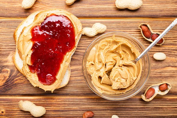Toastsandwich mit Erdnussbutter. Löffel und Glas Erdnussbutter, Marmelade und Erdnüsse zum Kochen des Frühstücks auf braunem Holzgrund. cremige Erdnusspaste flach liegend. — Stockfoto