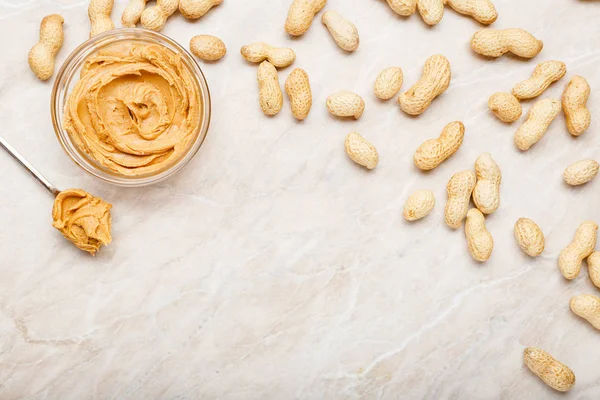 Erdnussbutter im Glasteller mit Erdnüssen in der Schale, Löffel mit Erdnussbutter. Cremige Erdnusspaste flach mit Platz für Text auf weißem Marmorhintergrund zum Kochen des Frühstücks — Stockfoto