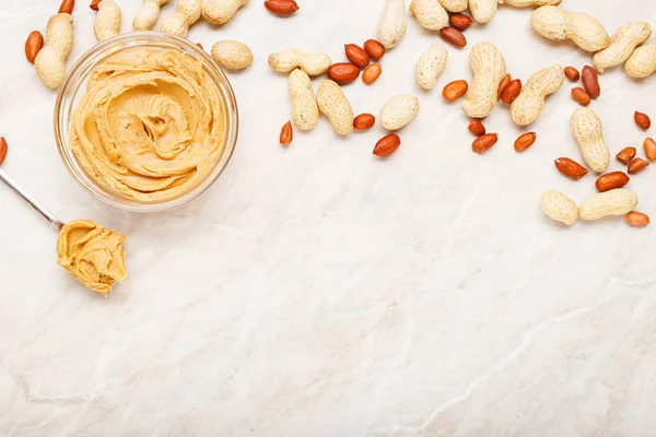 Erdnussbutter in Glasplatte mit Erdnüssen in Schale, geschälte Erdnüsse, Löffel mit Erdnussbutter. cremige Erdnusspaste flach mit Platz für Text auf weißem Marmorhintergrund zum Kochen des Frühstücks. — Stockfoto