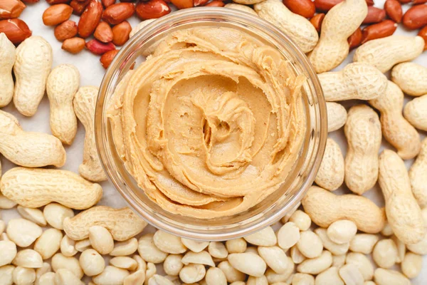 Creamy peanut paste, peanut butter in open glass jar in the center of peanuts food background. Peanuts in shell, peeled peanuts on white background . Vegan food concept. Creamy peanut paste flat lay.