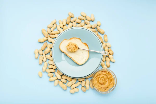 Toast with peanut butter, creamy peanut paste on blue plate on blue colored background. Served by peanut butter, peanuts in shell. Top view, flat lay. — 스톡 사진