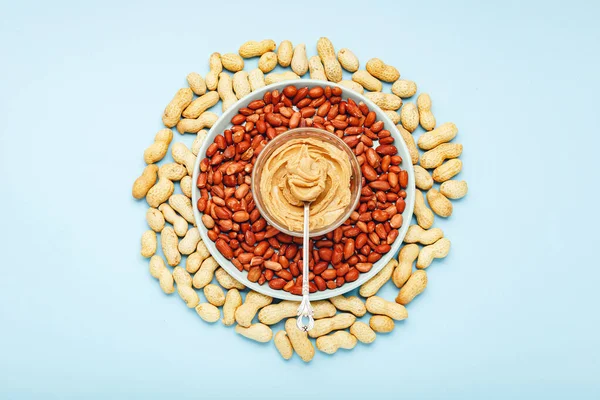 Creamy peanut paste in spoon on peanut butter on peanuts. Near the peanut butter lies peanuts in the shell and peeled peanuts. Minimalistic food flat lay on color blue background. Food background.