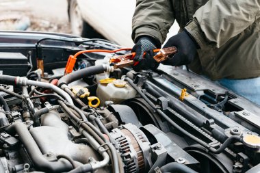 Açık havada akü kabloları kullanarak araba aküsünü elektrikle şarj eden makine mühendisi. Araba tamircisinin erkek ellerinde kırmızı ve siyah Jumper kabloları. Eldivenli adam araba tamirhanesinde çalışıyor..