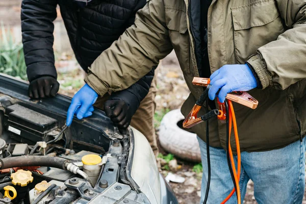 2 Mechanic engineers charging car battery with electricity using jumper cables outdoors. Red and black Jumper cables in male hands of car mechanic. Mans in gloves working in car repair service station — 스톡 사진