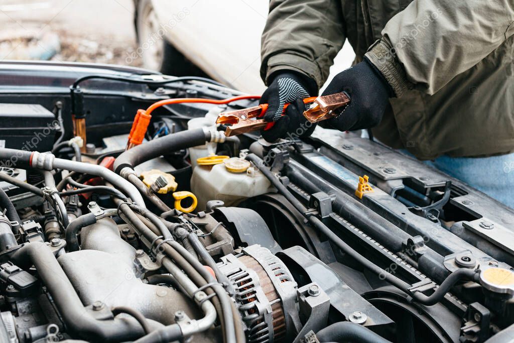 Mechanic engineer charging car battery with electricity using jumper cables outdoors. Red and black Jumper cables in male hands of car mechanic. Man in gloves working in car repair service station.