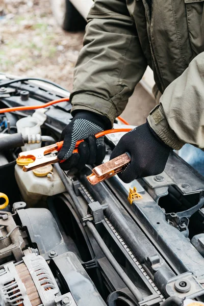 Mechanic engineer charging car battery with electricity using jumper cables outdoors. Red and black Jumper cables in male hands of car mechanic. Man in gloves working in car repair service station. — Stock Photo, Image