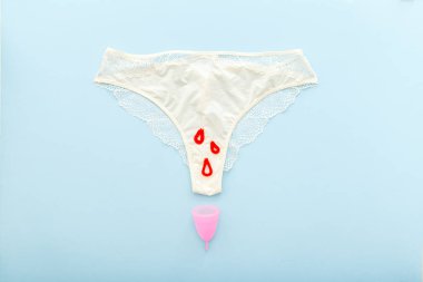 Pastel renkli mavi arka planda regl fincanı olan beyaz külot. İç çamaşırlarıyla, regl bardağıyla, kırmızı damlalarla yatıyordu. Regl, kadın hijyeni, kadın sağlığı, kritik günler, sıfır atık kavramı