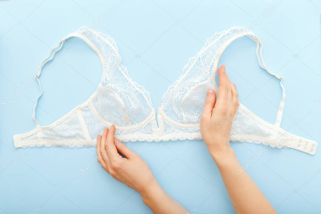 White Bra in female hands. Woman choosing holding white bra lingerie on blue background. Flat lay with beautiful romantic unlined elongated bralette. Sale, special offer, lingerie store concept.
