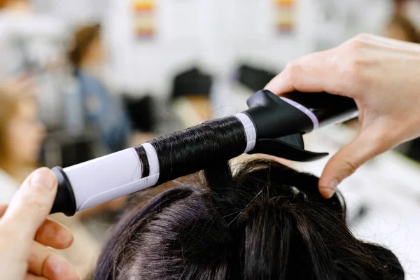 Hairstylist curling hair close up. Hairdresser making hairstyle to brown long hair woman in beauty salon. Professional hair care, Hairdressing services. Hair styling process