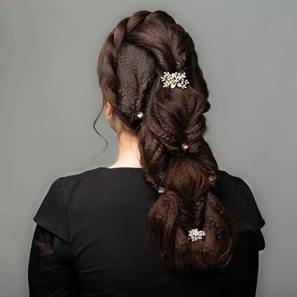 Weave, braid tail hairstyle. Hairstyle on brown hair woman with long hair on a gray background. Professional hairdressing services.Hair styling, making braid with hairpin