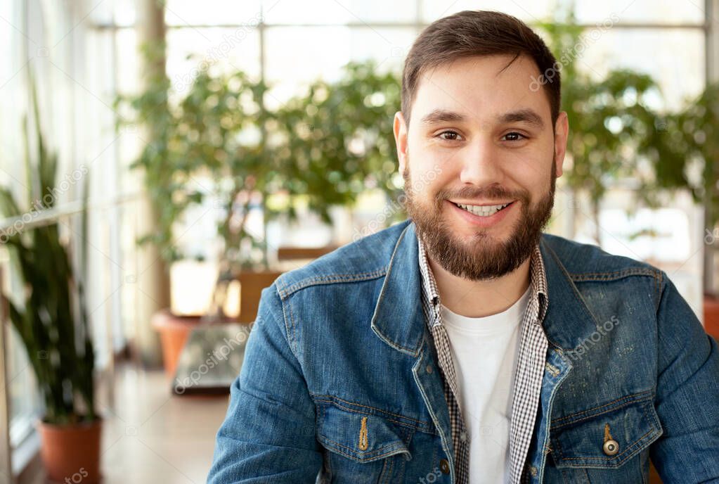 Portrait smiling handsome young man with beard in modern corporate environment near window. Smart man in office in casual wear, jean jacket. Attractive businessman freelancer smiling in cafe